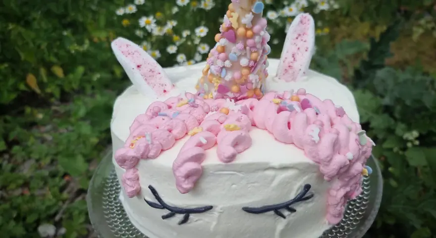 En förtrollande unicorn tårta med rosa glasstrut-liknande horn, dekorerad med strössel och blommor i bakgrunden.