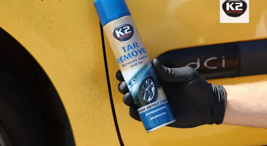 Rękawiczka z czarną gumą trzyma puszkę K2 Tar Remover, która skutecznie usuwa smołę z żółtego lakieru samochodu.