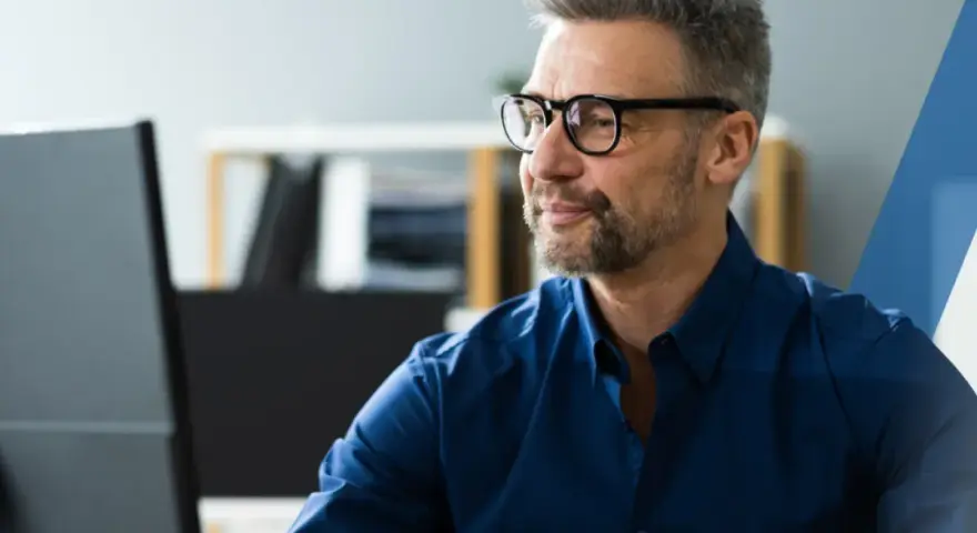 Hombre con gafas y barba gris, concentrado frente a su ordenador. Su camisa azul oscuro es un ejemplo de **art 16 et** en el vestuario profesional.