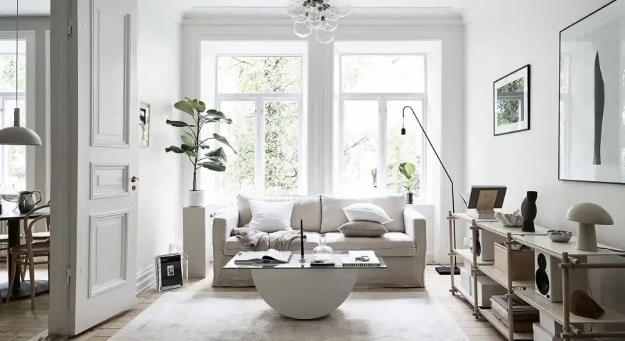 Salón con sofá beige, mesa de centro de cristal y alfombra clara. Decoración minimalista y plantas, creando un ambiente de feng shui.
