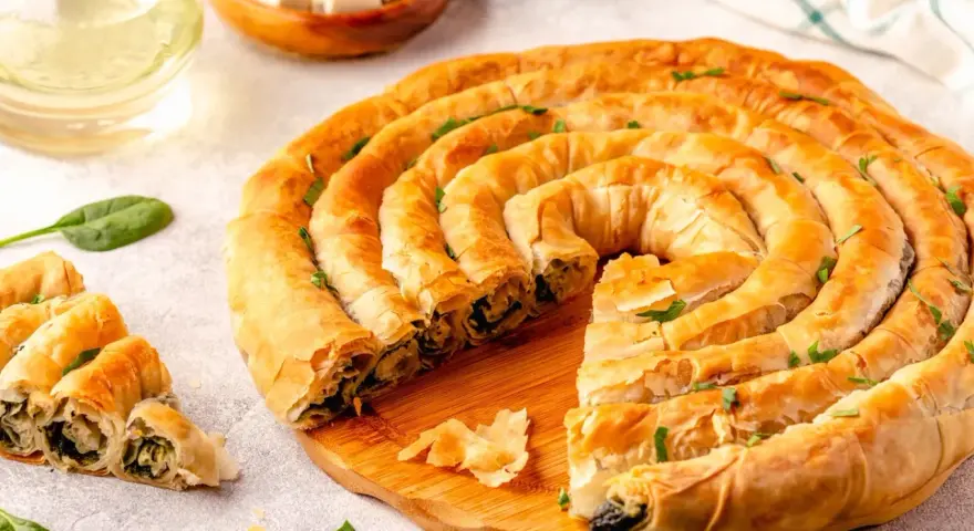 Złocisty borek ze szpinakiem, zwinięty w spiralę, kusi chrupiącym ciastem i aromatycznym nadzieniem. Obok kilka porcji i świeże liście szpinaku.