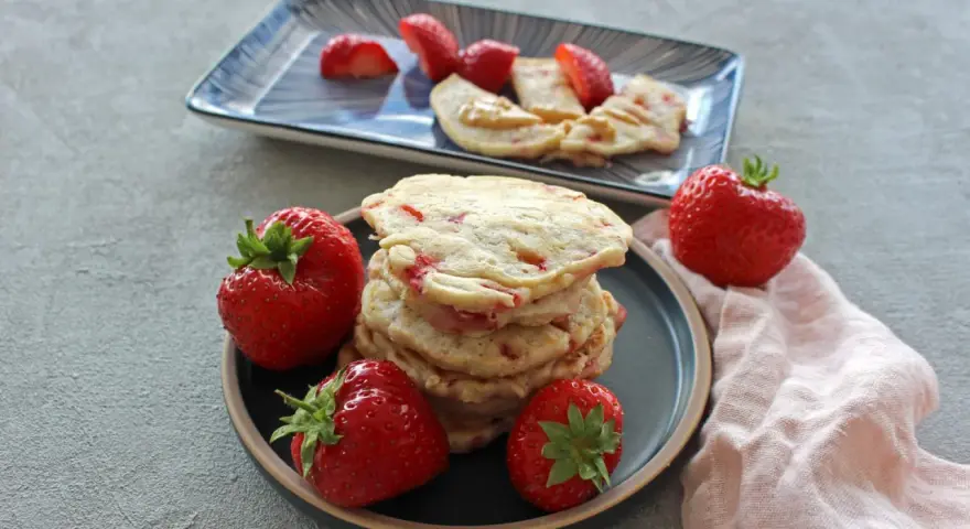 Ein Stapel Erdbeer-Pancakes mit frischen Erdbeeren auf einem Teller. Ein weiterer Teller im Hintergrund zeigt geschnittene Erdbeeren und Pancakes mit Erdnussbutter.