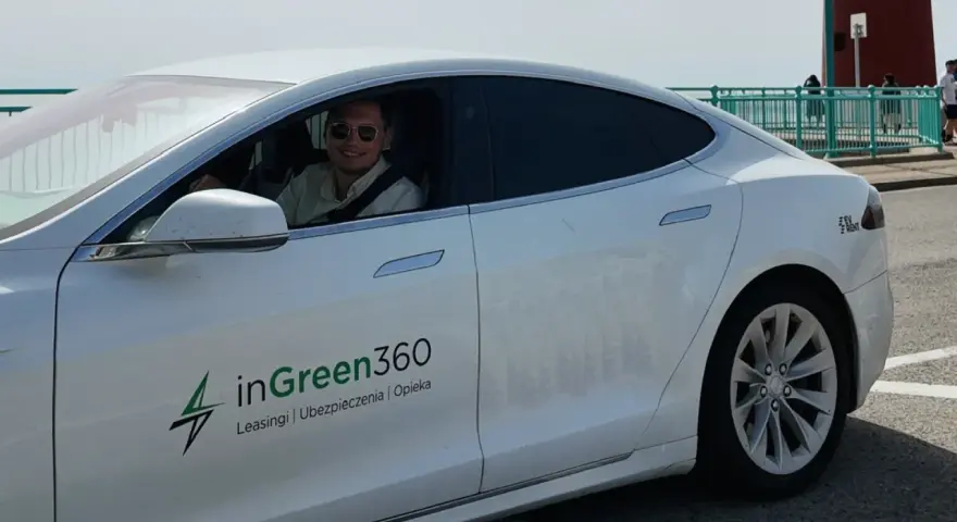 Uśmiechnięty mężczyzna w okularach przeciwsłonecznych siedzi za kierownicą białej Tesli z logo inGreen360.