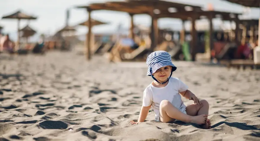 Uroczy maluch w pasiastym kapelusiku na plaży. Idealne miejsce, gdzie na wakacje z rocznym dzieckiem, by cieszyć się słońcem i piaskiem.