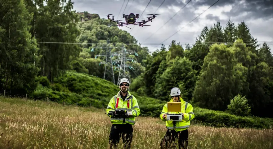 Two technicians in high-visibility jackets operate a power line drone in a grassy field, with a transmission tower and trees in the background.