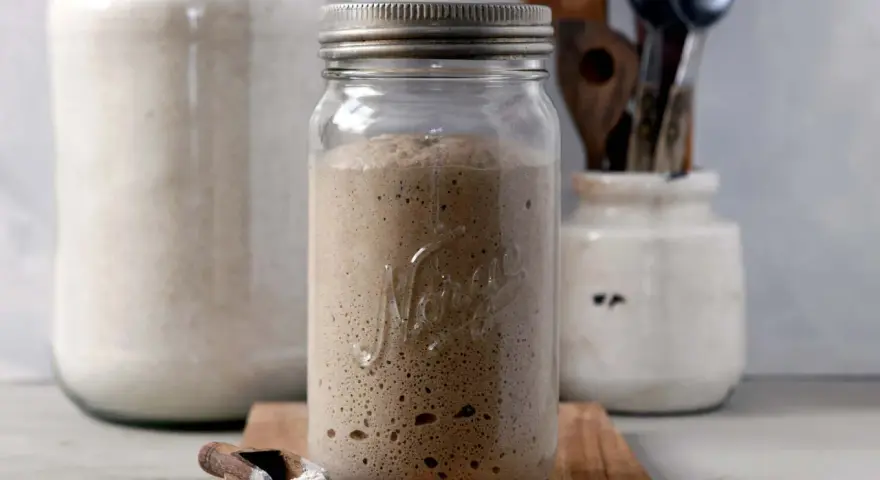 Blubbernde Sauerteig Kultur in einem Glas, bereit für das nächste Brot. Daneben ein Löffel mit Mehl und ein großer Behälter mit weiteren Zutaten.