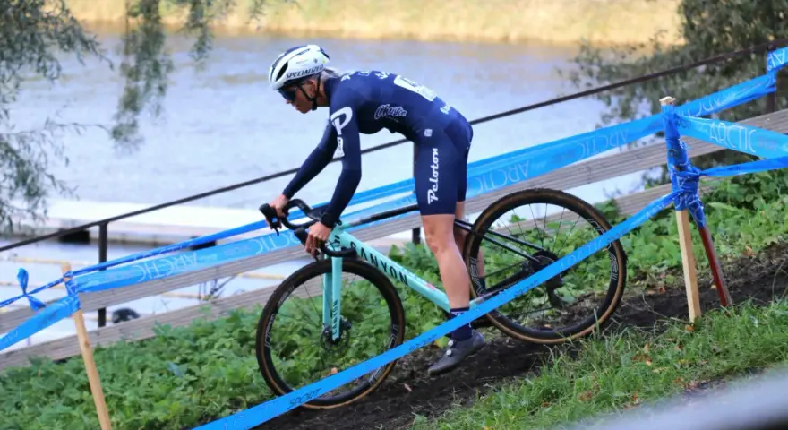 Cyclo cross racer in blue kit navigates a muddy course beside a lake, marked by blue tape.
