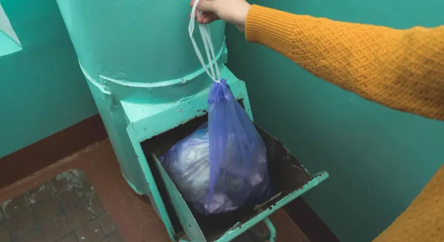 Ręka w żółtym swetrze wkłada worek ze śmieciami do metalowego zsypu w bloku.