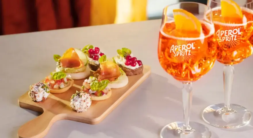 Zwei Aperol Spritz Gläser mit Orangenscheiben und eine Holzplatte mit verschiedenen Häppchen. Perfekt für einen Aperitif!