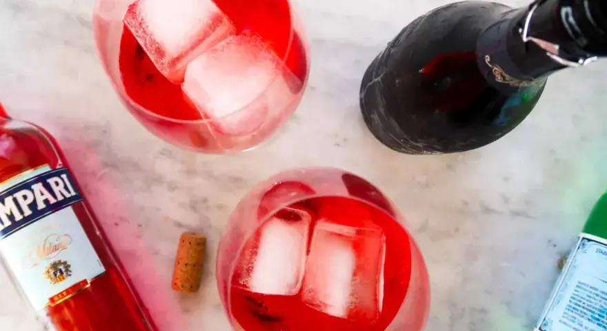 Dwa orzeźwiające Campari Spritz z lodem, butelka Campari i butelka prosecco na marmurowym blacie.
