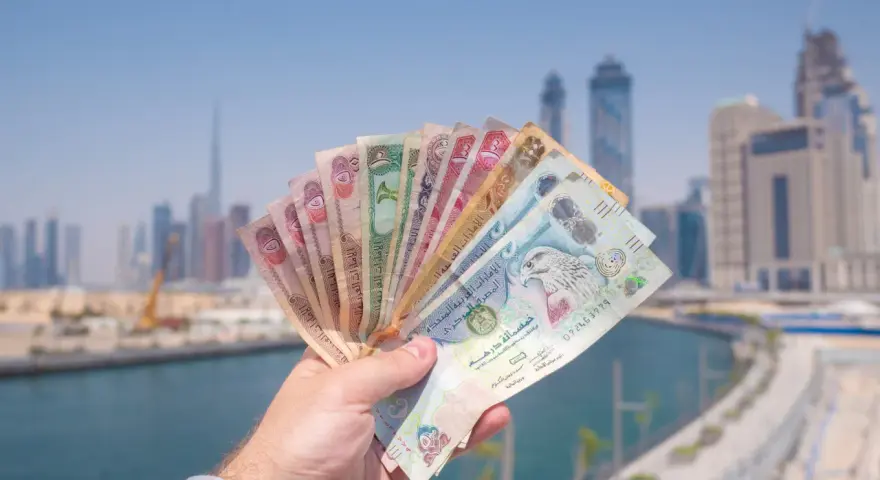 Dłoń trzymająca wachlarz banknotów dirhamów ZEA na tle panoramy Dubaju.