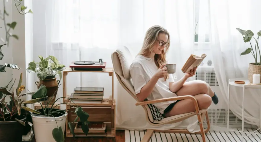 Mujer con gafas disfruta de un libro y una taza en su rincón de lectura, rodeada de plantas y luz natural.