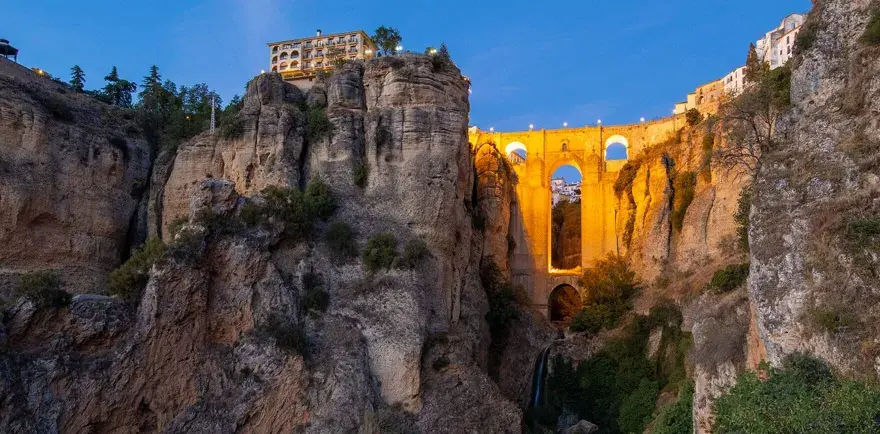 A dramatic **Ronda bridge view point** at dusk, with the illuminated bridge spanning a deep gorge and buildings perched on the cliff edge.