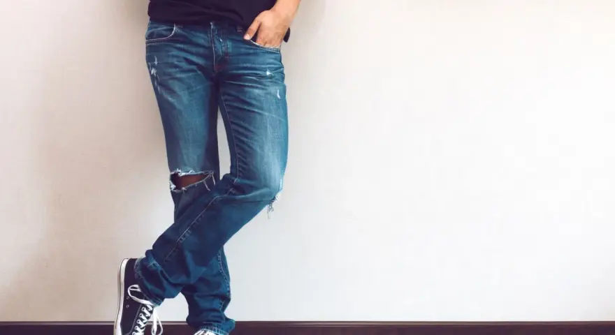 Mężczyzna w modnych jeansach z dziurami na kolanie, czarnych trampkach i czarnej koszulce, opiera się o ścianę.