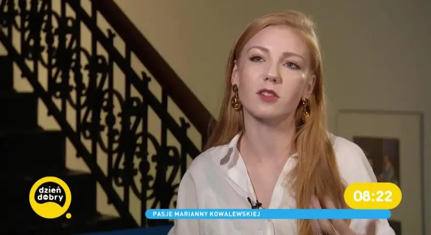 Marianna Kowalewska w białej koszuli, z rudymi włosami i złotymi kolczykami, opowiada o swoich pasjach na tle schodów.