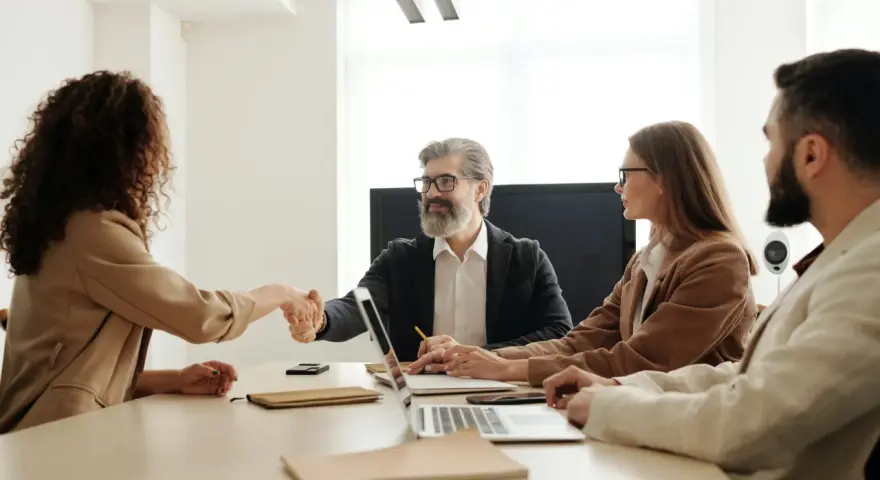 Uśmiechnięty mężczyzna ściska dłoń kandydatce, budując pozytywny employer branding. Przy stole siedzą też inne osoby.