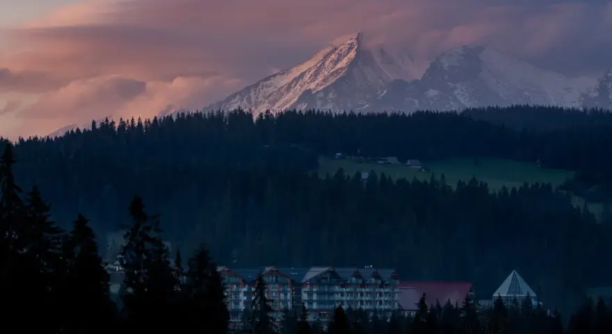 Górski krajobraz o zachodzie słońca z ośnieżonymi szczytami, lasem i budynkami hotelowymi.