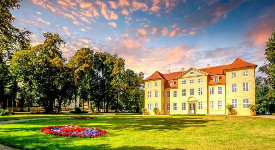 Gelbes Schloss mit roten Dächern, umgeben von grünen Bäumen und Blumenbeeten. Ein malerischer Anblick auf der Liebesinsel Mirow.