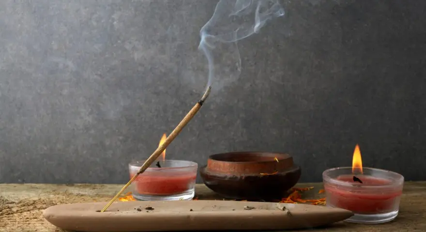 Un incienso humeante se posa sobre un soporte de arcilla, rodeado de velas rojas y un cuenco de madera. Perfectos inciensos para limpiar energías y crear un ambiente de paz.