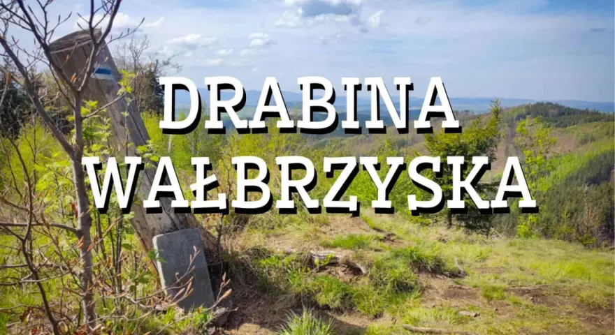 Widok na górskie krajobrazy z napisem "Drabina Wałbrzyska". Stary drewniany słup z niebieskim znakiem szlaku stoi na zboczu porośniętym trawą i młodymi drzewkami.