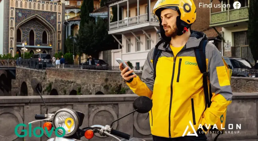 Kurier Glovo sprawdza telefon, gotowy do pracy w blokach czasowych. Na skuterze widnieje logo.