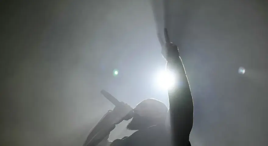 Un rappeur, micro en main, lève un doigt en signe de paix. Lumière intense, fumée, ambiance d'un exposé sur le rap.