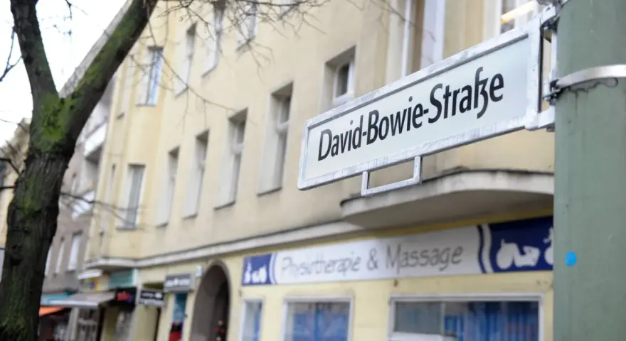 Straßenschild "David-Bowie-Straße" in Berlin. Ein Baum und ein Gebäude mit Geschäften im Hintergrund.