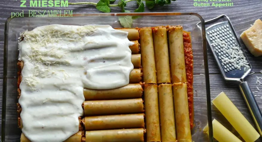 Cannelloni z mięsem mielonym i sosem beszamelowym w naczyniu żaroodpornym, gotowe do pieczenia. Obok tarka i kawałek parmezanu.