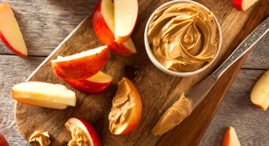 Saftige Apfelspalten mit cremiger Erdnussbutter auf einem Holzbrett. Ein Messer mit Erdnussbutter liegt daneben.