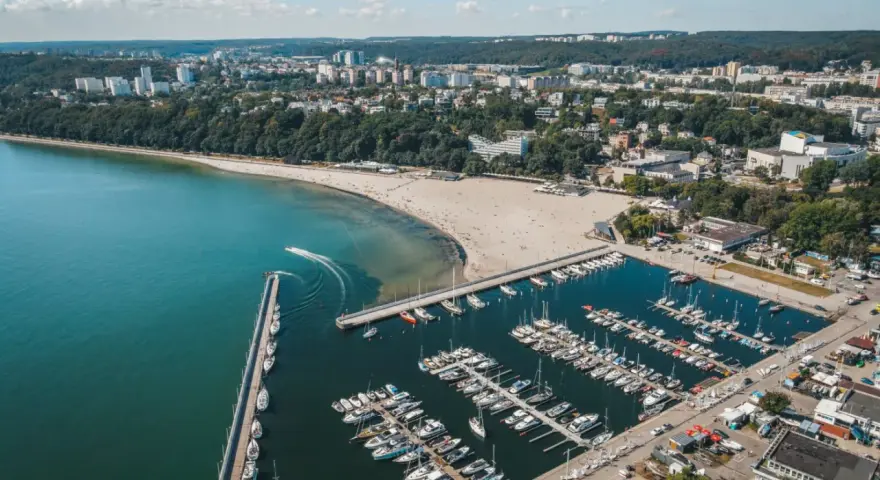Widok z lotu ptaka na plażę miejską w Gdyni, z zatoką pełną jachtów i łodzi, oraz miastem w tle.
