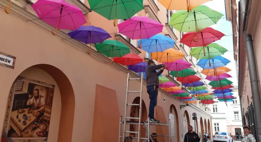 Kolorowe parasolki tworzą tęczowy dach nad uliczką w Tarnowie. Mężczyzna na drabinie dokańcza instalację.