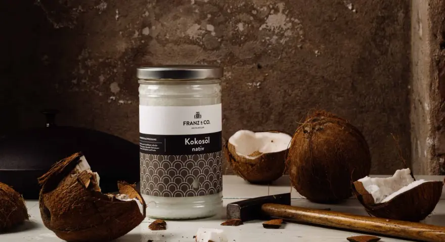Ein Glas Kokosöl nativ steht neben zerbrochenen Kokosnüssen und einem Hammer. So wird aus der Frucht das wertvolle Öl gewonnen.