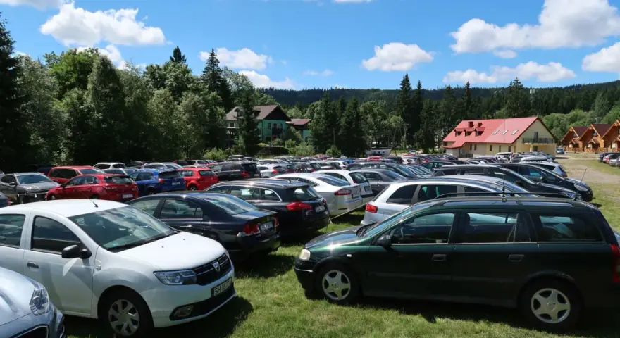 Tłum samochodów na parkingu przy Karłowie Szczeliniec. Słoneczny dzień, chmury na niebie, otoczenie lasu i budynków.