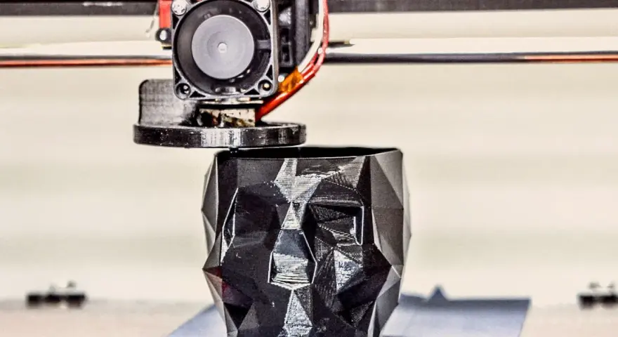 A 3D printer nozzle extrudes filament to create a faceted black skull planter, showcasing innovative 3D printing examples.