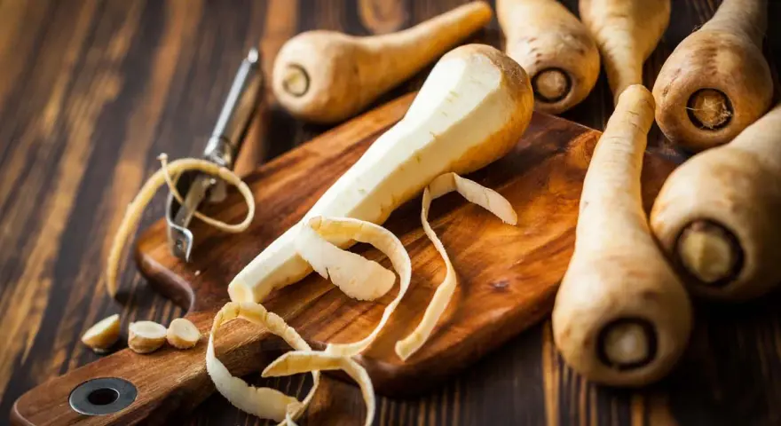 La pastinaca pianta, parzialmente pelata su un tagliere, circondata da altre radici e un pelapatate.