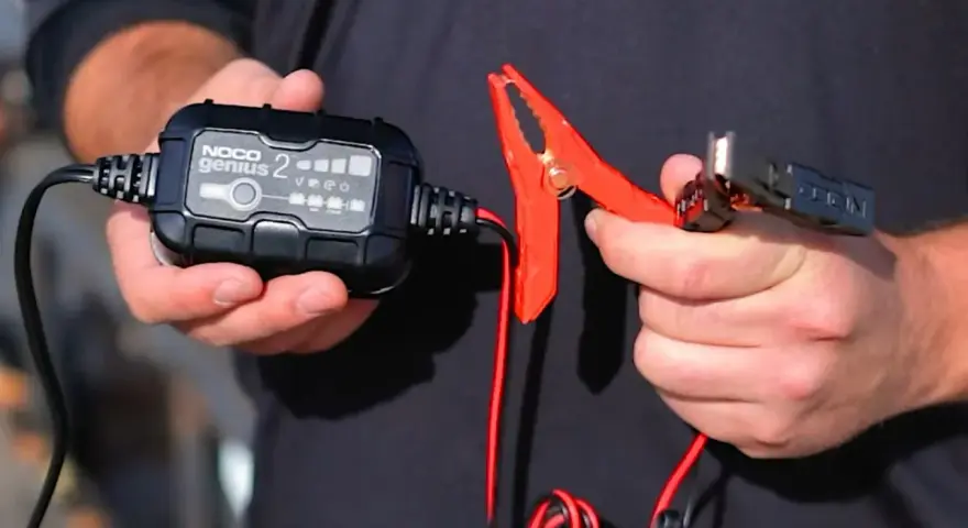 A person holds a NOCO Genius 2 trickle charger and its red clamp. This charger is designed to prevent damage, so no, a trickle charger can't ruin a battery when used correctly.