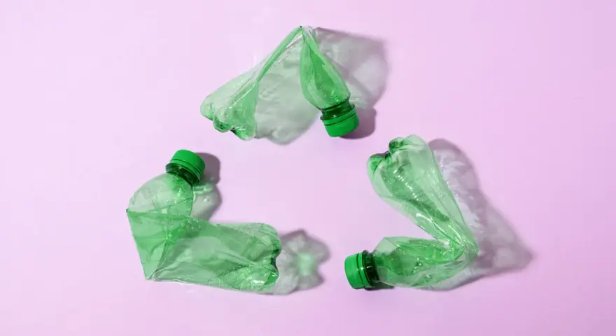 Trzy zielone, zgniecione butelki tworzą symbol recyklingu na różowym tle.