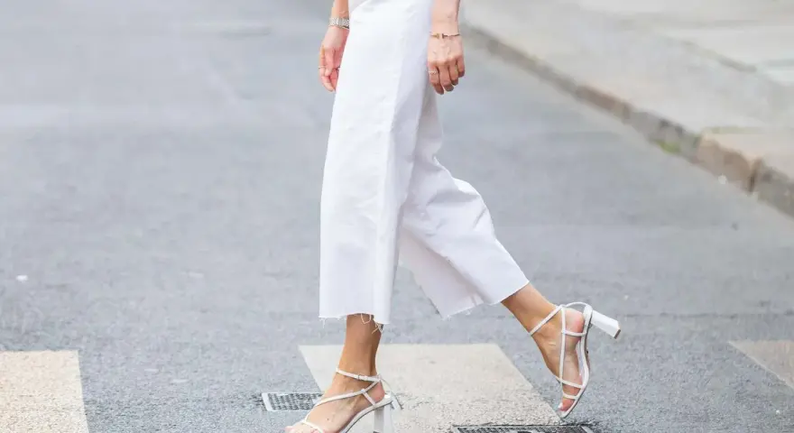 Mujer cruza la calle con un look comunión: pantalón blanco de corte ancho y sandalias de tiras.