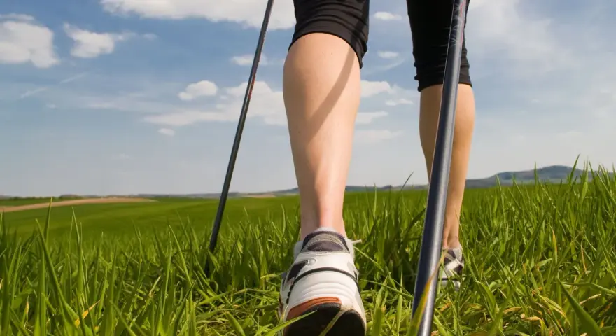 Gambe che praticano nordic walking tecnica su un prato verde sotto un cielo azzurro con nuvole bianche.