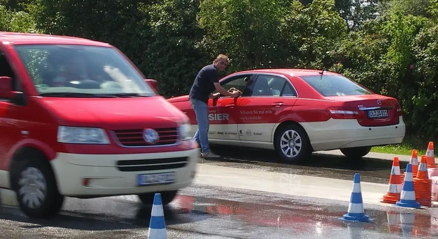 Mann reinigt Taxi. Ein roter Transporter fährt vorbei. Verkehrskegel markieren den Bereich. Möglicherweise eine Schulung der Berufsgenossenschaft Taxi.