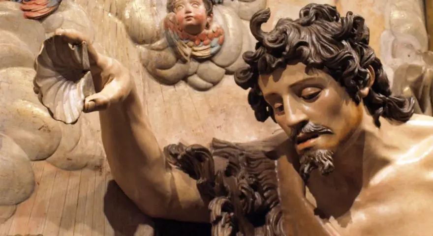 Detalle de escultura barroca: figura masculina con barba y cabello rizado sostiene una concha. Ángeles infantiles observan. Maestría de escultores del barroco.