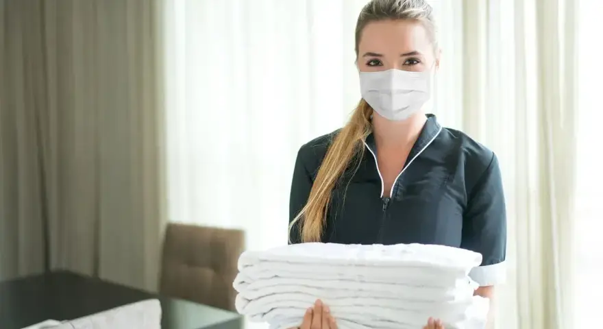Pracownica hotelu w maseczce niesie stos czystych ręczników. Odpowiedź na pytanie: czy w hotelach są ręczniki - tak!