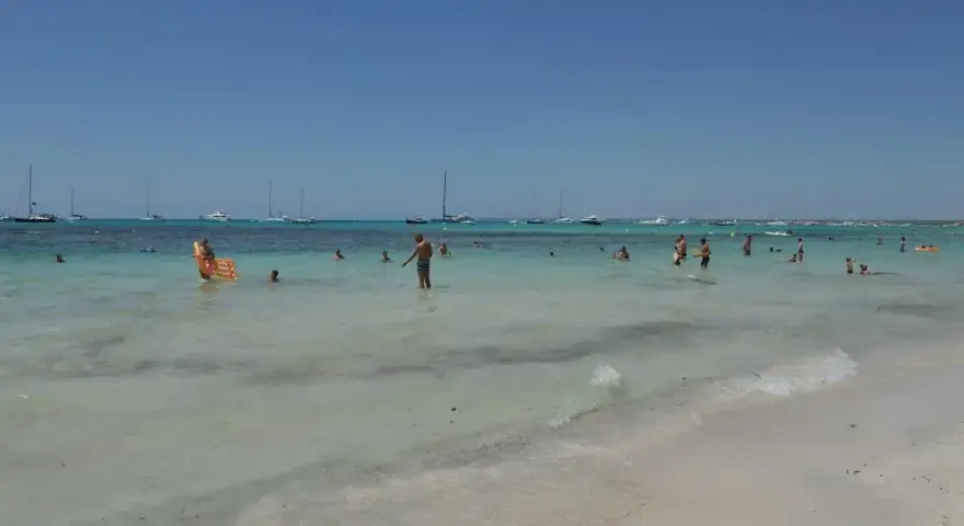 Słoneczny dzień na plaży Es Trenc. Ludzie kąpią się w turkusowej wodzie, a na horyzoncie widać żaglówki.