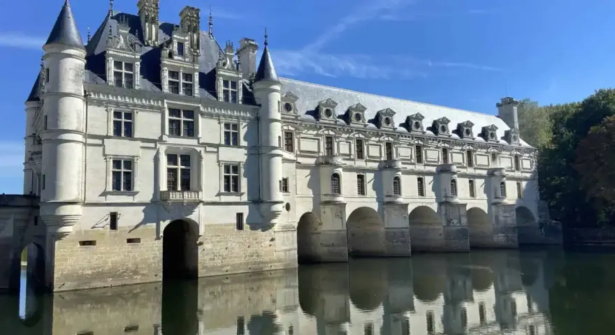 The stunning Chateau de Chenonceau, a white stone castle, gracefully spans the River Cher, its reflection shimmering on the water's surface under a clear blue sky.