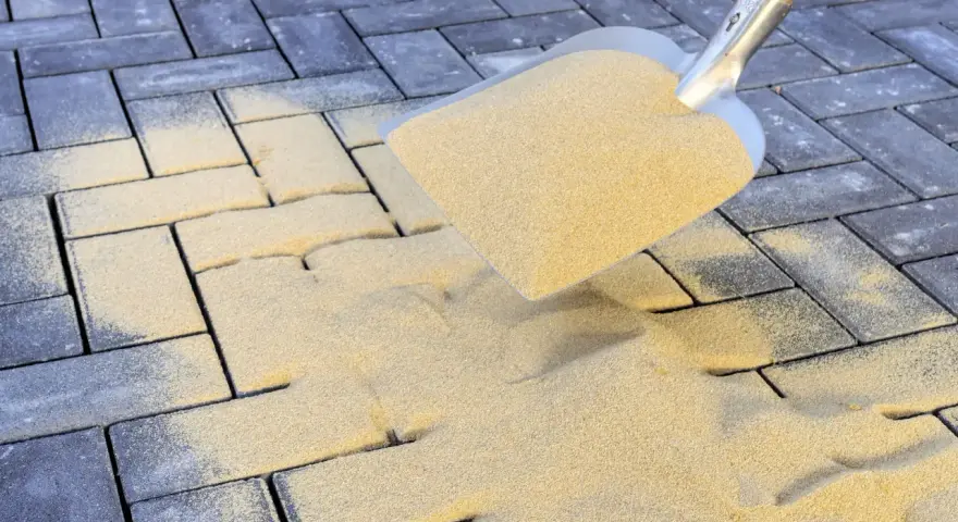 Schaufel mit Sand zum Terrassenplatten verfugen. Sand rieselt auf graue Pflastersteine.