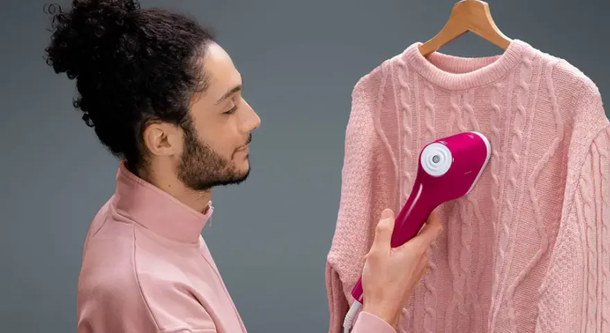 Man using a pink handheld steamer on a pink cable-knit sweater hanging on a wooden hanger.