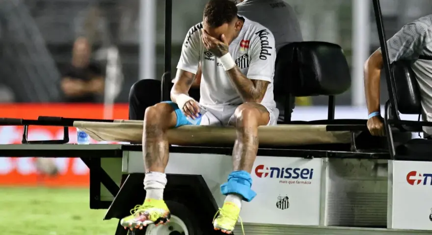 Neymar siedzi na wózku golfowym, zakrywając twarz dłońmi. Jego kontuzja jest widoczna, a zawodnik wygląda na zrozpaczonego.