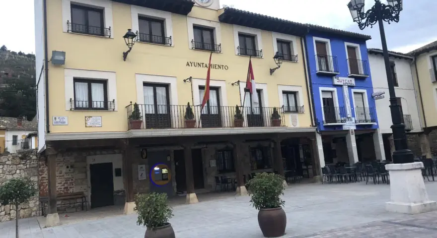 El Ayuntamiento de Tajuña, con su balcón y reloj, preside la plaza. Edificios de colores y farolas completan la escena.