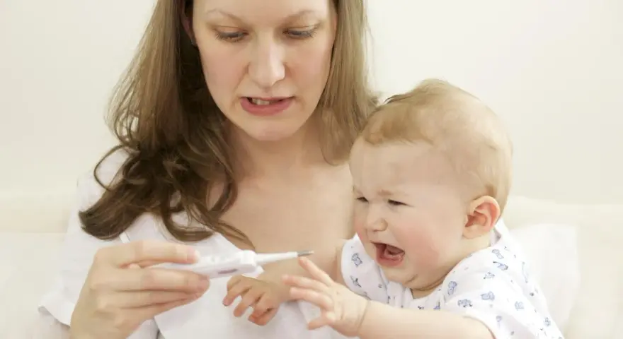 Zmartwiona mama mierzy temperaturę płaczącemu dziecku, które ma gorączkę po szczepieniu.