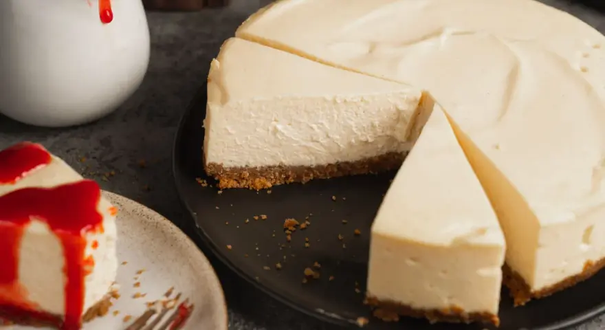 Ein cremiger New York Cheesecake, das Original-Rezept, mit einem Stück auf einem Teller, beträufelt mit roter Soße.